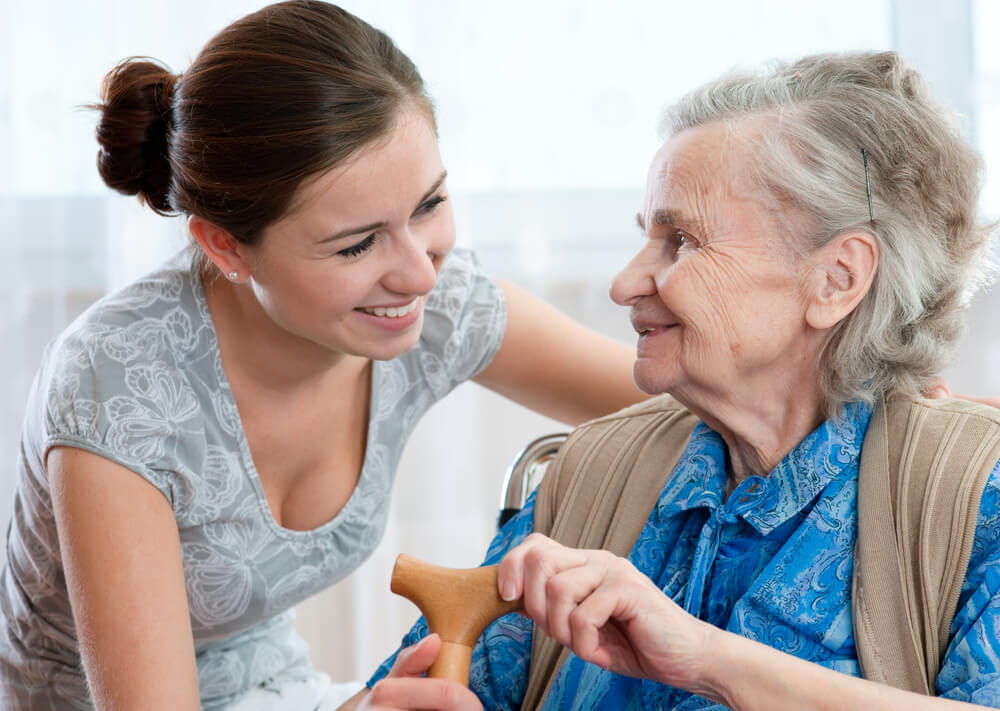 Young woman with brown hair interacts with an older woman sitting down holding a cane.